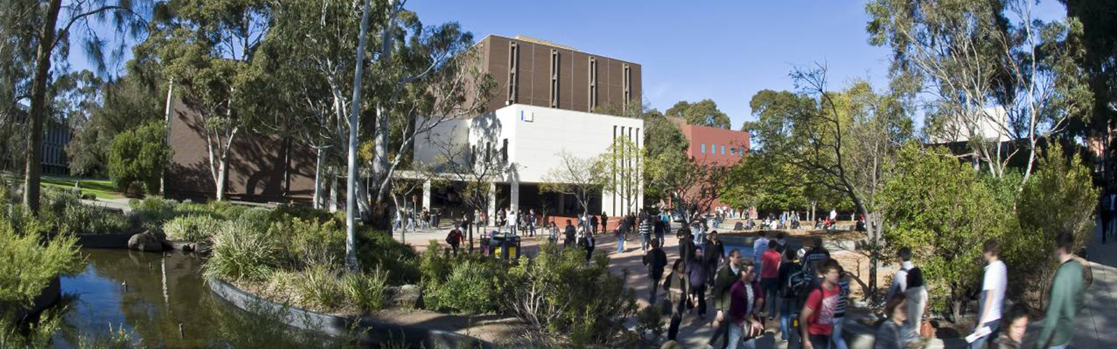 Students are congregating outside one of the university buildings. Greenery surrounds the buildings. 